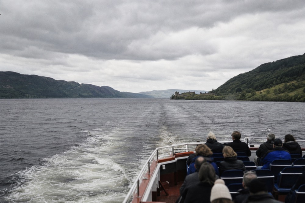 Open-deck sightseeing boat cruising down Loch Ness towards Urquhart Castle in clear daylight
