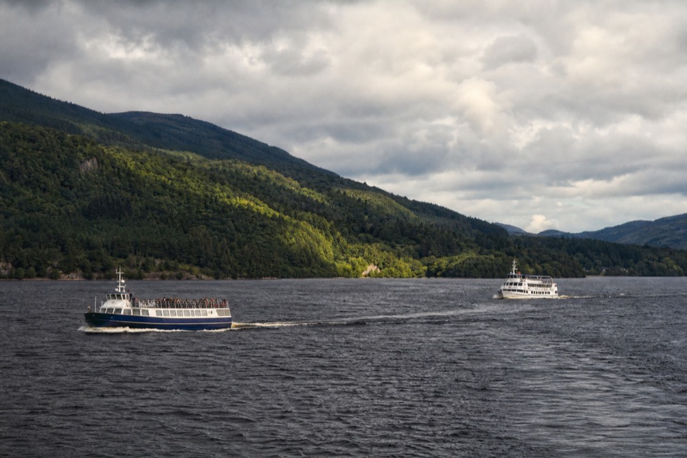 A white passenger cruise boat cutting across the dark water of Loch Ness with wooded hills rising behind