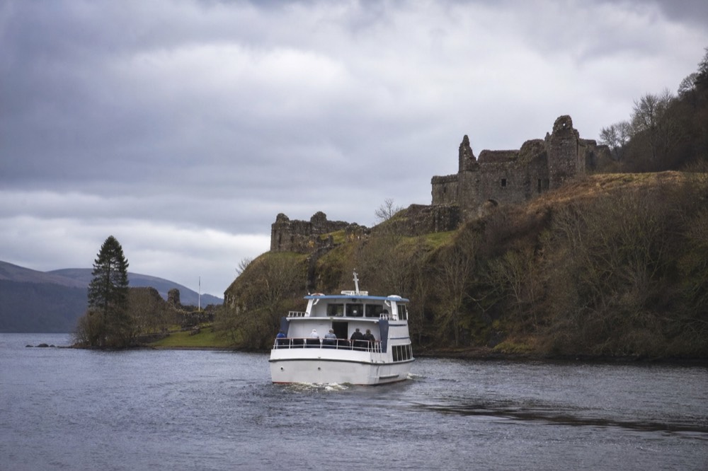Urquhart Castle on its headland seen from a passenger boat approaching across the dark water of Loch Ness