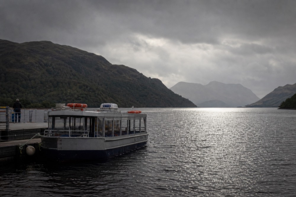 A cruise boat under way on Loch Ness in golden evening light with wooded ridges rising behind it