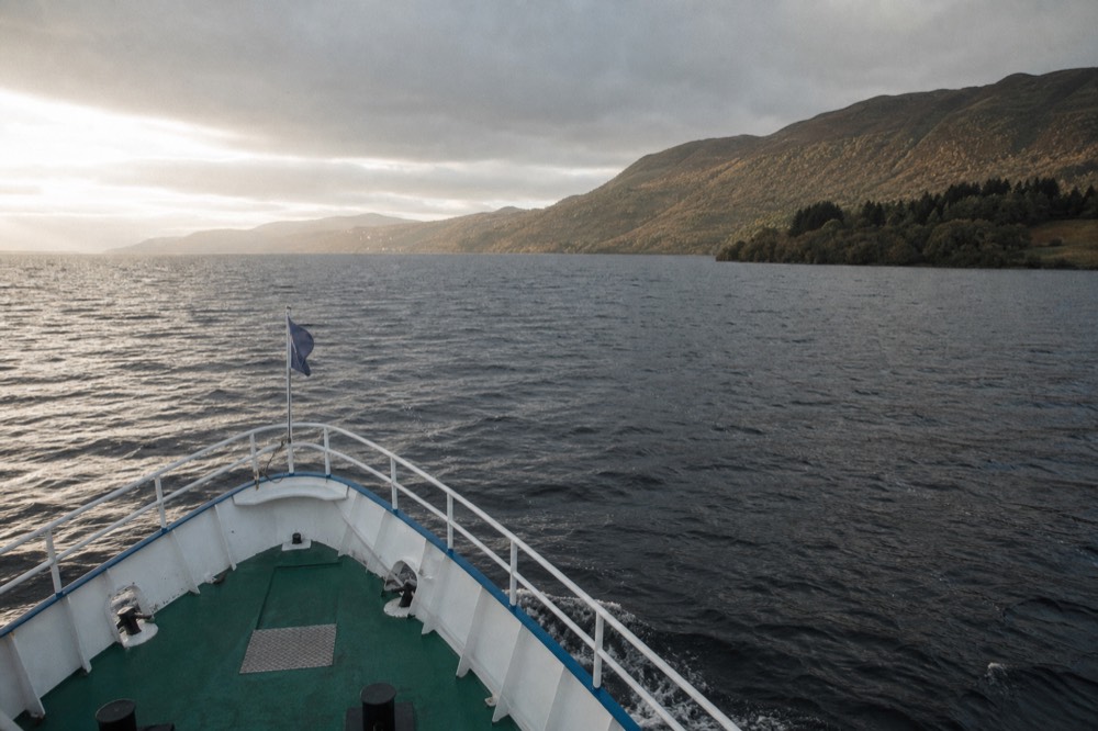 A cruise boat cutting a wake across the dark water of Loch Ness with steep Highland hills behind